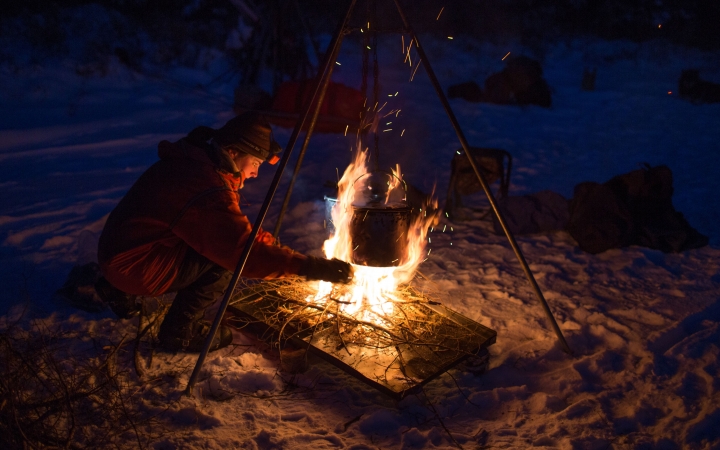 a person tends to a campfire in the snow. There is a tripod holding a pot over the fire. 